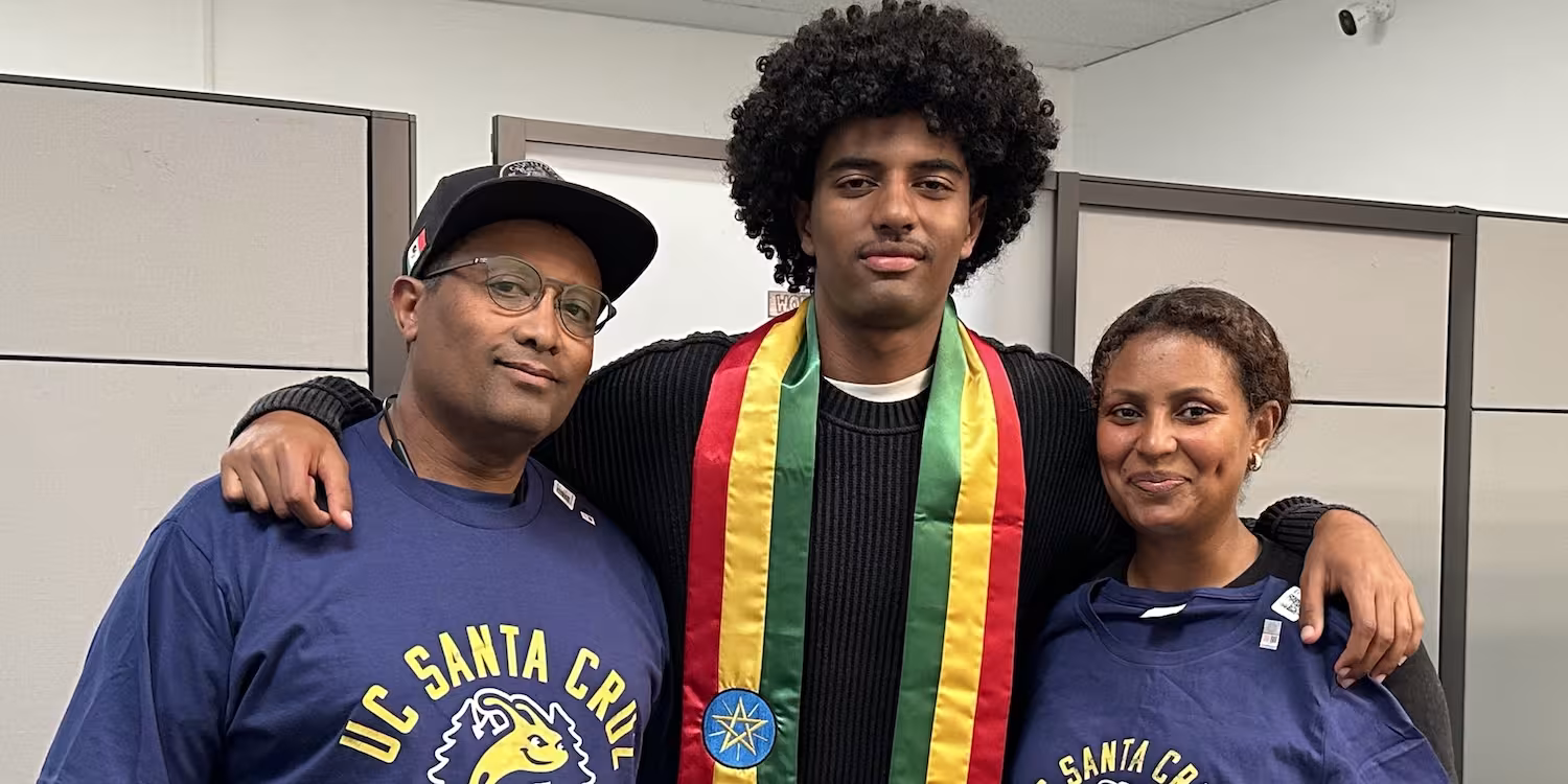 A graduate wearing a sash standing with a man and woman after a graduation ceremony.