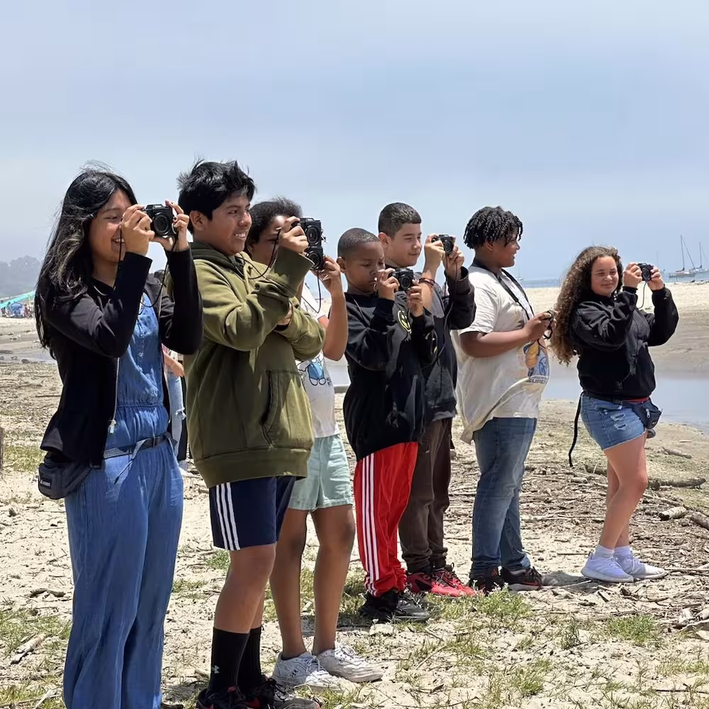 Students looking through binoculars on a beach during a summer learning activity.