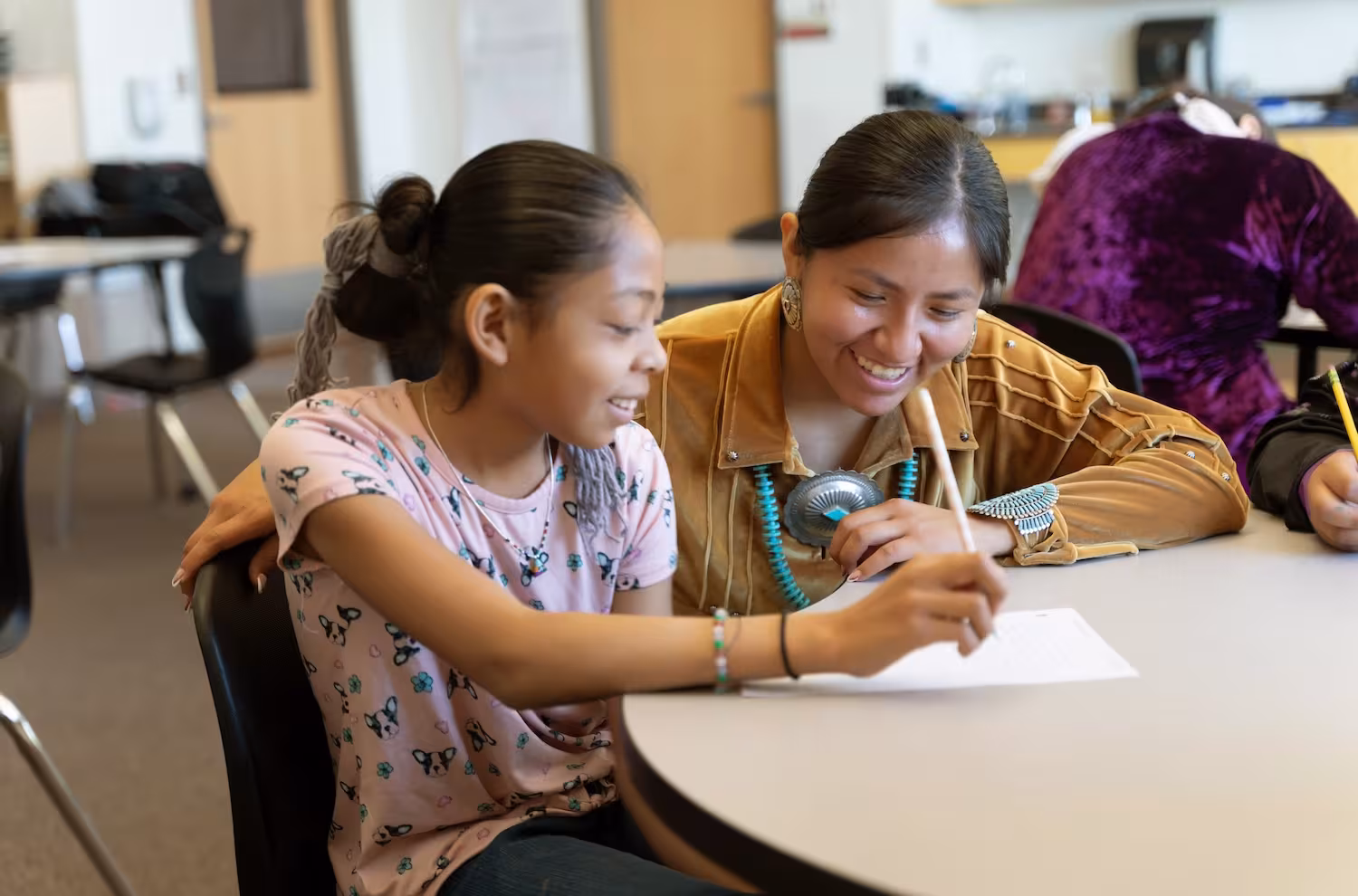 A student works on a writing activity with guidance from a smiling mentor in a classroom.